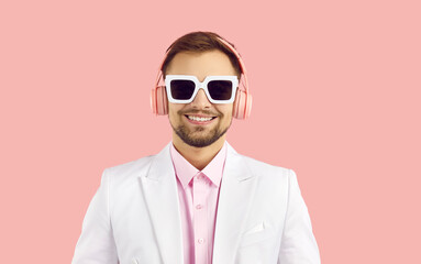 Portrait of smiling young man in modern stereo headphones isolated on pink background. Close up of cheerful guy in white jacket and sunglasses listening to music with modern wireless headphones.