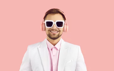 Foto op Canvas Portrait of smiling young man in modern stereo headphones isolated on pink background. Close up of cheerful guy in white jacket and sunglasses listening to music with modern wireless headphones. © Studio Romantic