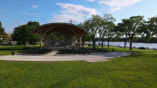 aerial view of park bandshell with covering along with Mississippi River. Bright blue sky with fluffy white clouds and green grass. Walking paths all around. 