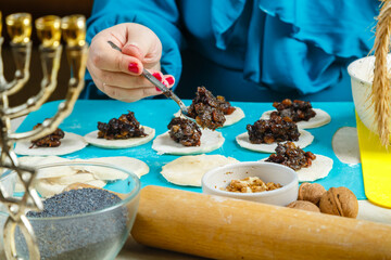 The hands of a Jewish woman laying poppy seed filling on the dough for gomentash biscuits for the Purim holiday in the foreground of the menorah.