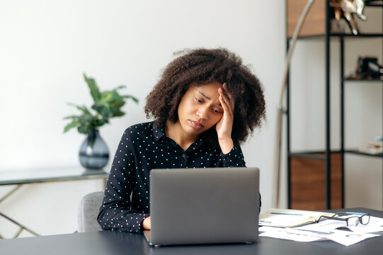 Upset depressed young adult african american woman, top manager, office worker, director of the company, sits at the desktop in front of the laptop, looks at the screen in disappointment, got bad news