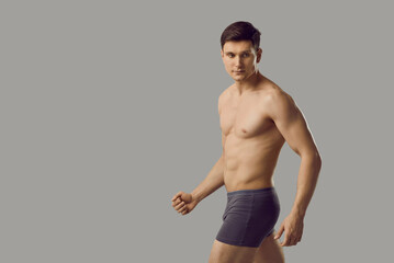 Young man with perfect athletic body in underwear and with naked torso walks on gray background....