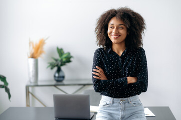 Portrait successful curly-haired confident beautiful young adult afro american businesswoman or broker, stands near desk in office dressed in stylish clothes, arms crossed, looking to the side, smile © Kateryna