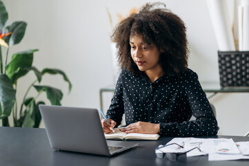 Young adult African American business woman in the office. Attractive confident smart woman top manager or trader, sits in front of a laptop, looks the screen, makes working notes in a notebook