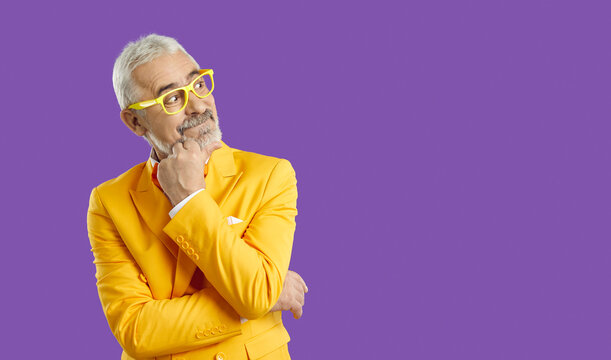 Portrait of funny happy curious bearded senior man wearing bright yellow suit and eyeglasses, with hand on chin thinking about what he sees on blank solid purple copyspace background on right side