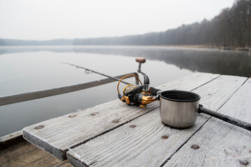 Fishing rod, spinning reel and cup of hot coffee on the wooden background pier river bank. Sunrise....