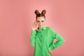 Young stylish woman in green shirt and round glasses with two bun hairstyles on pink background.