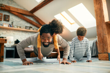 Happy black father has fun while doing push-ups with his kids at home.