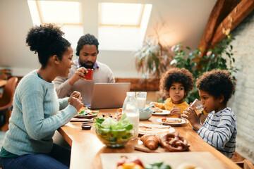 Happy black mother talks to her children during breakfast at dining table while father is working...