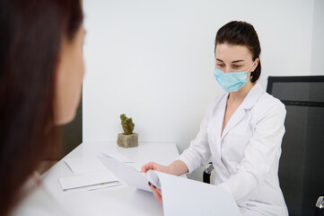 Woman at a doctor's appointment in a white office. Confident general practice cosmetologist beautician wearing medical protective mask reading medical documentation during examination her patient