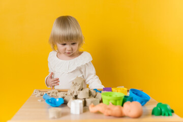 A beautiful girl in a white dress is standing near table with kinetic sand, plastic molds and other...