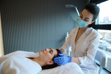 Naklejka premium Relaxed elegant Caucasian woman enjoying a professional massage on her head done by a massage therapist while lying on a couch in a wellness spa center.