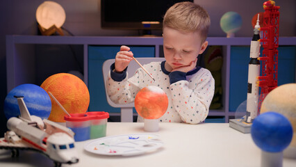 Blond boy paints planet solar system Mars with colorful paints sitting home table in evening,...