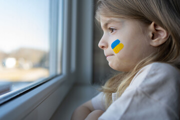 a crying child, looking out the window, with painted on her cheek the yellow-blue colours of the...
