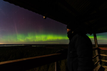 Aurora borealis, The Northern lights at the lake Usma and forest, Latvia. Wooden watching tower. Aerial view. The man is watching the northern lights.