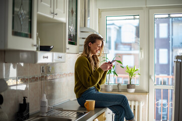 Woman standing in the kitchen and drinking cup of tea while using smartphone