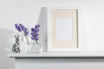 White hyacinth in a large porcelain bowl, books, figurines of hares and a bird, an empty photo frame are on the fireplace against the white wall. Layout.