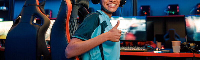 Happy African female cybersport gamer wearing headset showing approval sign while sitting on gaming chair in internet cafe