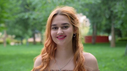 Beautiful girl with red hair laughs while looking at the camera in nature in summer