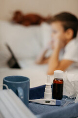Bottle with antipyretic syrup, nasal spray and thermometer on tray. Little boy in bed in foreground...
