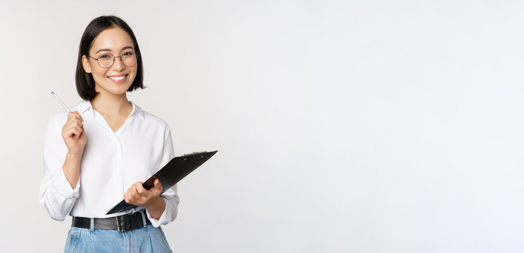 Smiling Young Asian Woman Taking Notes With Pen On Clipboard, Looking Happy, Standing Against White Background