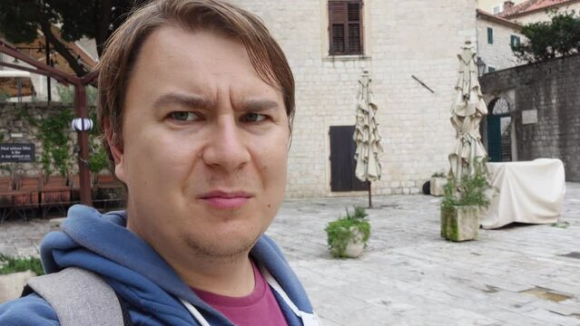 Young man walks through deserted streets of old town decorated with stone and tells something emotionally, actively gesticulating and looking up. Travel blogger is streaming a new video