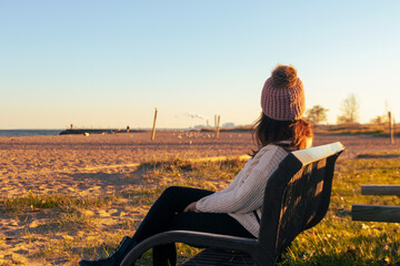 A female wearing a puffy winter had sitting on a bench during a sunset and looking away into the distance.