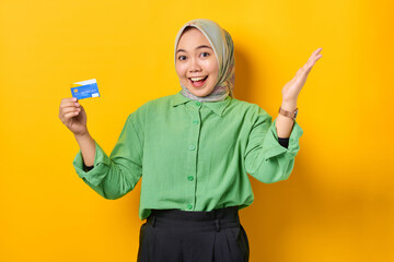 Excited young Asian woman in a green shirt holding credit card while spreading hand on yellow background