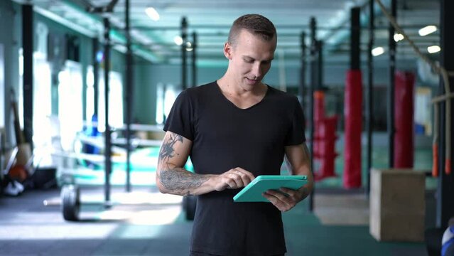 Tattooed handsome Caucasian man standing in gym surfing Internet on tablet and smiling looking at camera. Positive sportsman posing indoors in slow motion after training. Workout and satisfaction