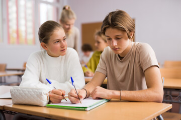 Teenage boy and girl listening to teacher and writing exercises in notebook at lesson in secondary...