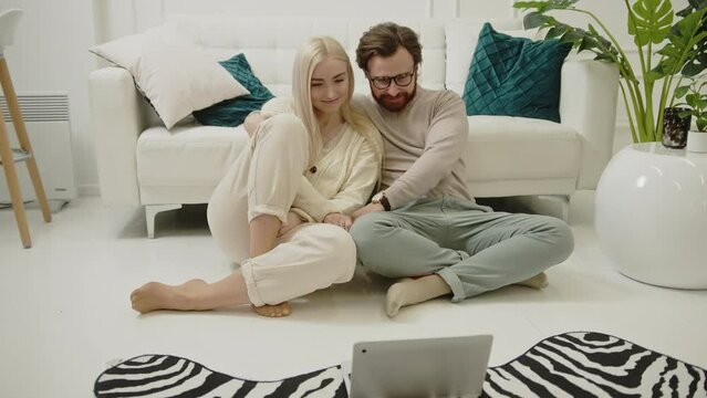 European blonde long-haired beauty sitting on the floor with her affectionate partner and watching videos on their laptop. High quality 4k footage