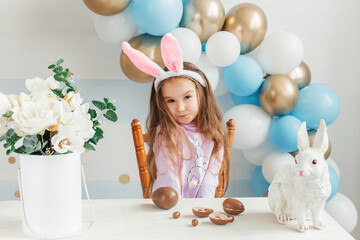 Little cute girl in easter bunny ears sits at the table and plays with easter chocolate eggs. Happy easter
