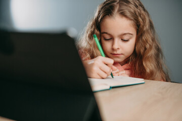 Caucasian little girl with tablet pc computer writing to notebook at home. Online education. Kid learn online learning school. Quarantine, pandemic.