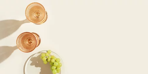 Foto auf Alu-Dibond White wine glasses peach color glass and grapes fruit on beige background with dark shadow, glare from sun. Minimal summer rest concept. Dry wine in colored glassware goblets style. Creative © yrabota