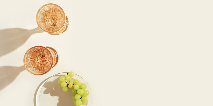 White Wine Glasses Peach Color Glass And Grapes Fruit On Beige Background With Dark Shadow, Glare From Sun. Minimal Summer Rest Concept. Dry Wine In Colored Glassware Goblets Style. Creative