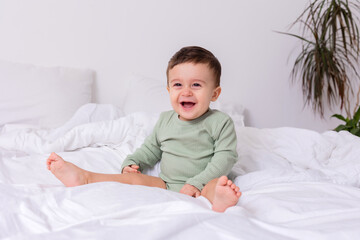 laughing baby in a green cotton bodysuit and bare feet is sitting on a bed spread out