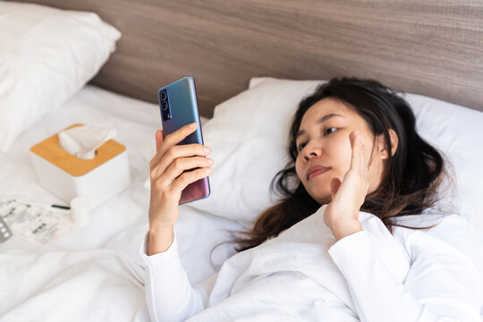 Sick young Asian woman sleep on bed in bedroom at home and video call with doctor or her family on mobile phone. Cold, flu, home isolation during coronavirus pandemic  concept. Closeup