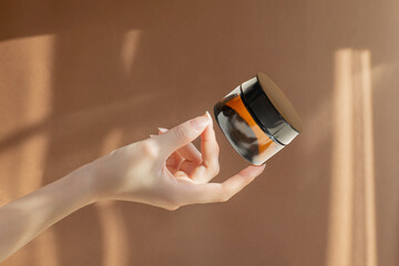 Woman's hand holds jar of moisturizing cream made of amber glass on brown background. Bottle...