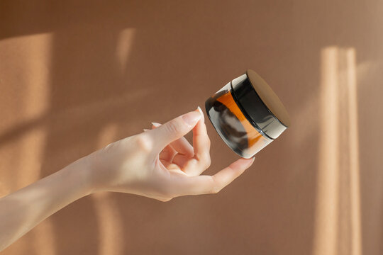 Woman's hand holds jar of moisturizing cream made of amber glass on brown background. Bottle without marking with anti-aging mask, cosmetic. Concept of skin care