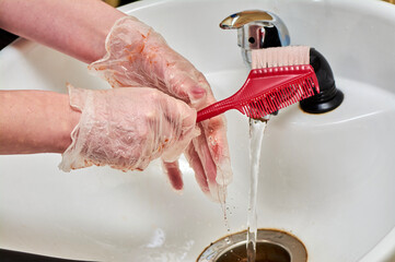 Women's hands in hair coloring gloves wash a comb under a stream of water in a white sink