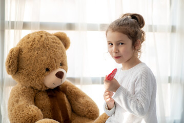 A little girl plays with a big teddy bear. She is dressed in a white jumper. Childhood. Lifestile.