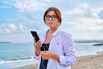 Middle age sad serious woman in headphones with smartphone outdoor.