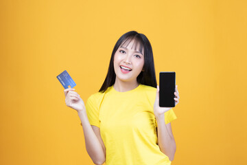 Portrait Asian happy young girl smiling cheerful and showing plastic credit card while holding mobile phone isolated on yellow background
