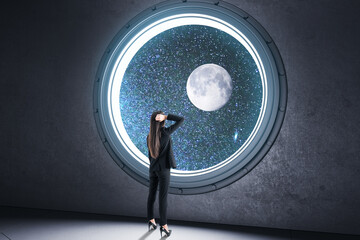 Back view of young businesswoman looking out of round illuminator with starry sky cosmos and planet view in concrete interior. Future and creativity concept.