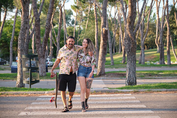 Fototapeta premium Young cool tattooed couple walking together with a longboard in a park outdoors looking at camera.