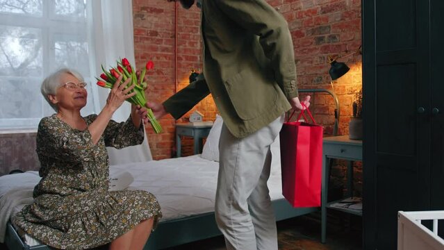pleasantly surprised elderly grey-haired pensioner woman receiving a bouquet and a gift from her middle-aged son Mother's Day . High quality 4k footage