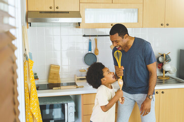 Happy African American Father with Cute son dancing and holding kitchen appliance microphones singing while cooking in the kitchen at home.