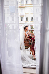 A beautiful young woman in exquisite lingerie stands on the balcony of a multi-storey building and holds a large bouquet of beautiful flowers in her hands. Morning of the bride on the wedding day.