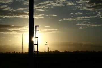 Lìnea de torres de alta tensiòn en silueta al atardecer con el sol cayendo en el horizonte, forma...