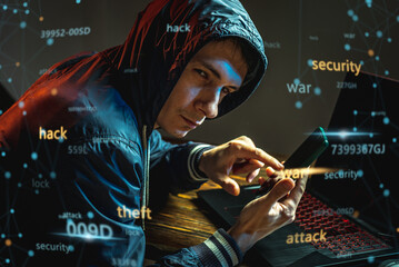 Hacker in a hood with a phone is typing on a laptop keyboard in a dark room. Cybercrime fraud and...